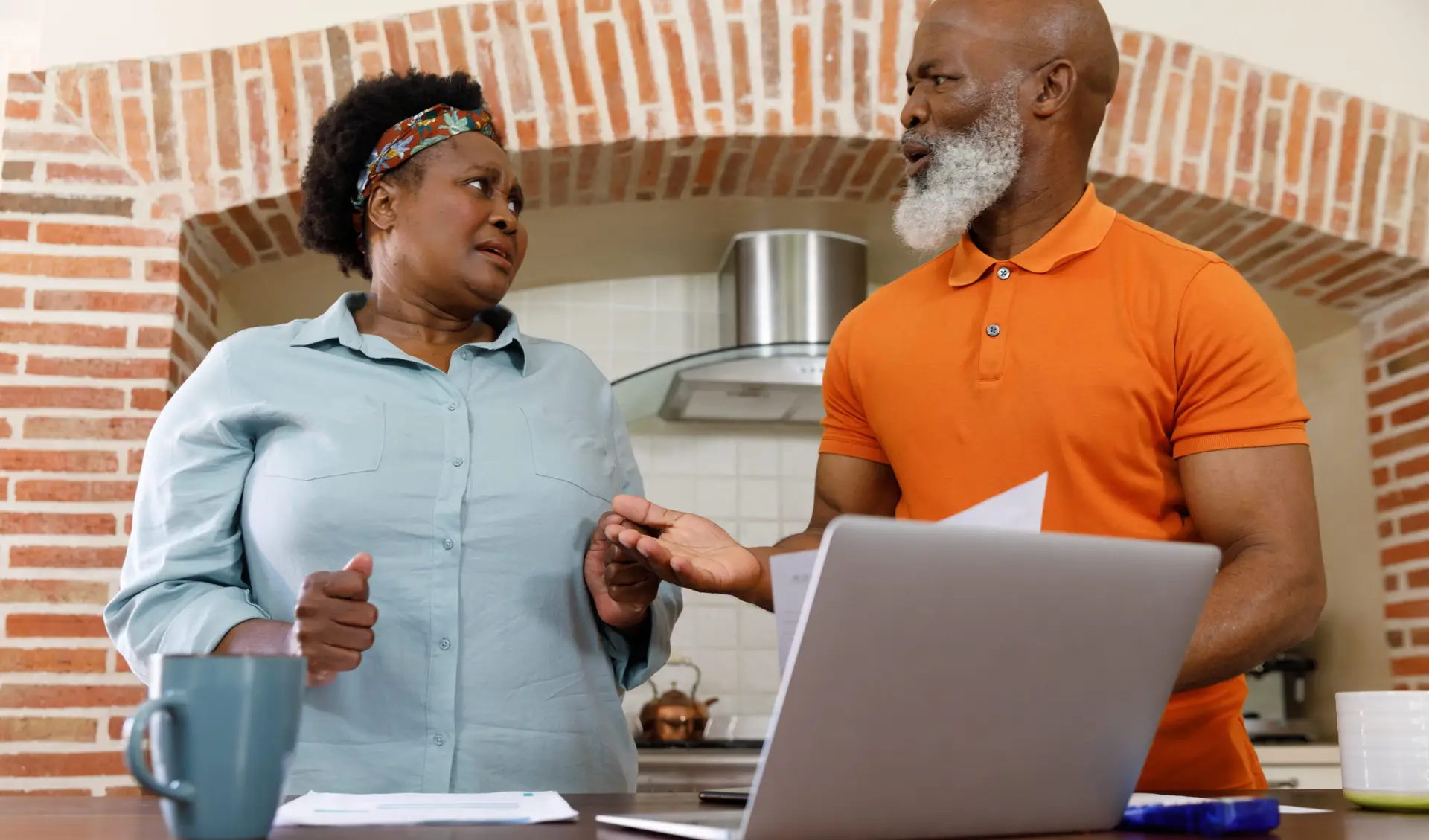 An older couple stands at a kitchen counter with a laptop and paperwork, looking concerned as they discuss their finances.